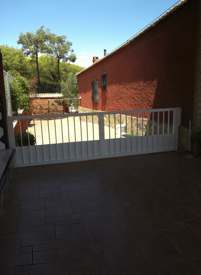 Puerta de aluminio de dos hojas abatibles en color blanco con barrotes y chapa inferior para impedir paso de animales.